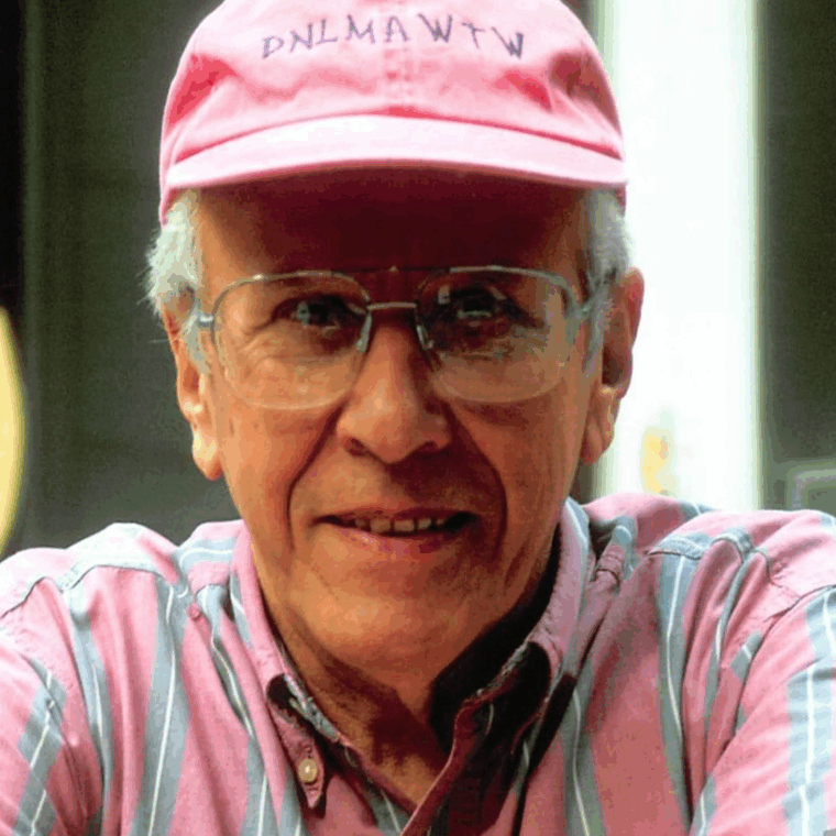 An older man wearing a pink cap with embroidered letters, glasses, and a pink striped shirt, smiling at the camera in a brightly lit setting.