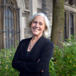 A smiling older woman with short gray hair stands outdoors in front of a stone building and green bushes, wearing a black cardigan and a necklace, with her arms crossed.