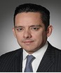 A man with short dark hair is dressed in a pinstripe suit and white dress shirt with a gray tie. He has a neutral expression and is posing against a dark gradient background.