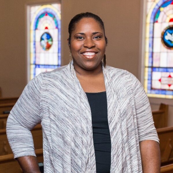 A person smiling warmly indoors, standing near wooden benches in a room with stained glass windows. They are wearing a gray cardigan over a black top.