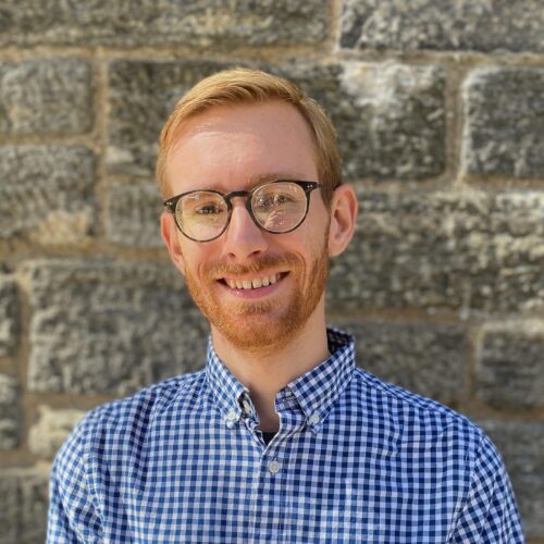 A person with short red hair, a beard, and glasses is smiling in front of a stone wall. He is wearing a blue and white checkered shirt.