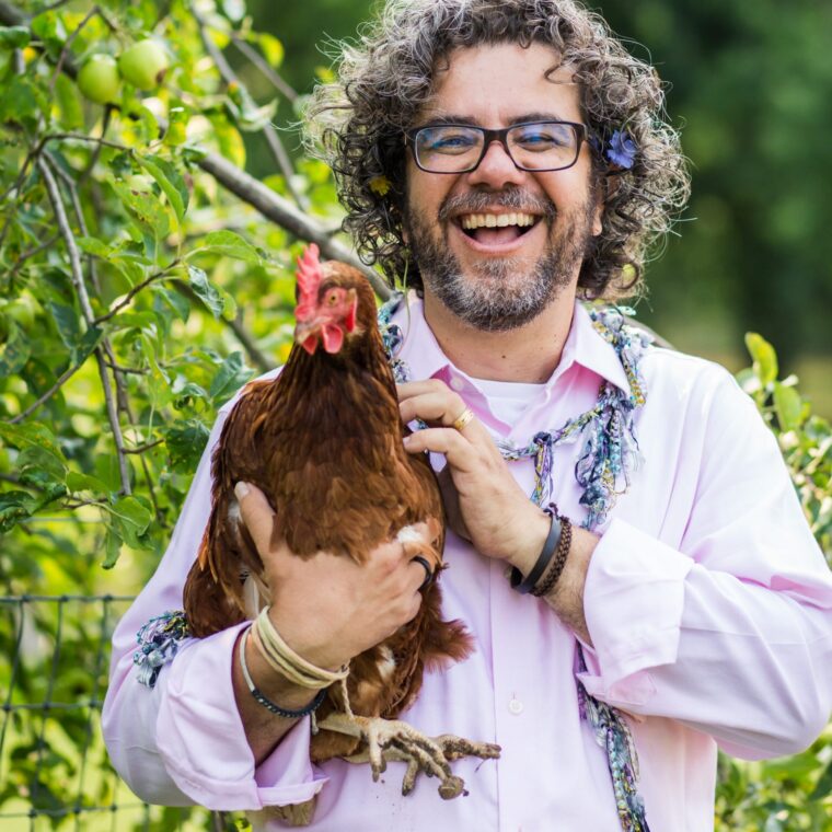 A smiling man with curly hair and glasses, wearing a light pink shirt, stands outdoors holding a brown chicken. Green foliage and apples are visible in the background.