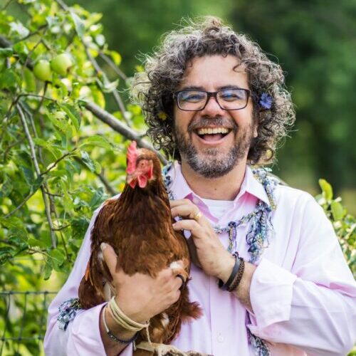 A smiling man with curly hair and glasses, wearing a light pink shirt, stands outdoors holding a brown chicken. Green foliage and apples are visible in the background.