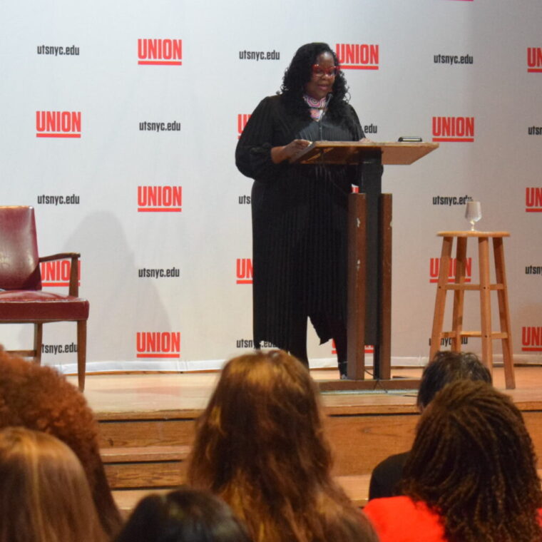 A woman speaks at a podium on a stage in front of an audience. Behind her is a backdrop with “Union” and “utsnyc.edu” repeated. Two empty chairs and a stool are also on the stage.