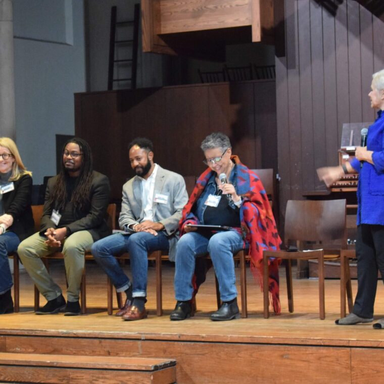 Five people are seated on a stage, with four sitting in chairs and one standing. They appear to be engaged in a discussion. The setting seems to be an auditorium with wooden decor.