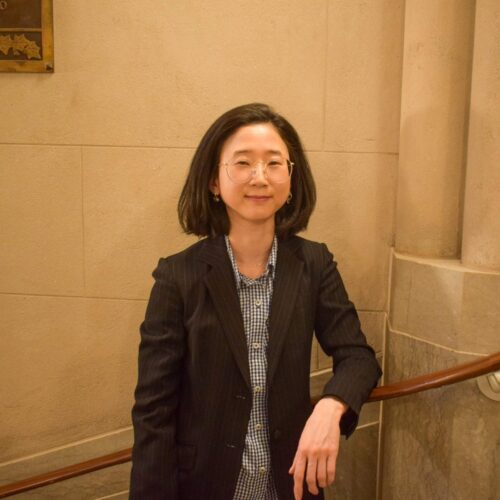 A person with shoulder-length hair and glasses is standing confidently in front of a staircase with large stone columns. They are wearing a dark blazer over a patterned shirt and are smiling softly.