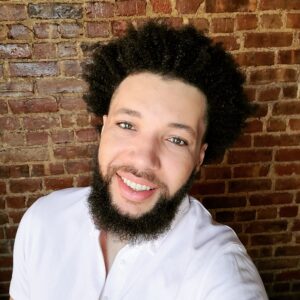 A person with curly hair and a beard smiles at the camera while wearing a white shirt. They stand in front of a textured brick wall.