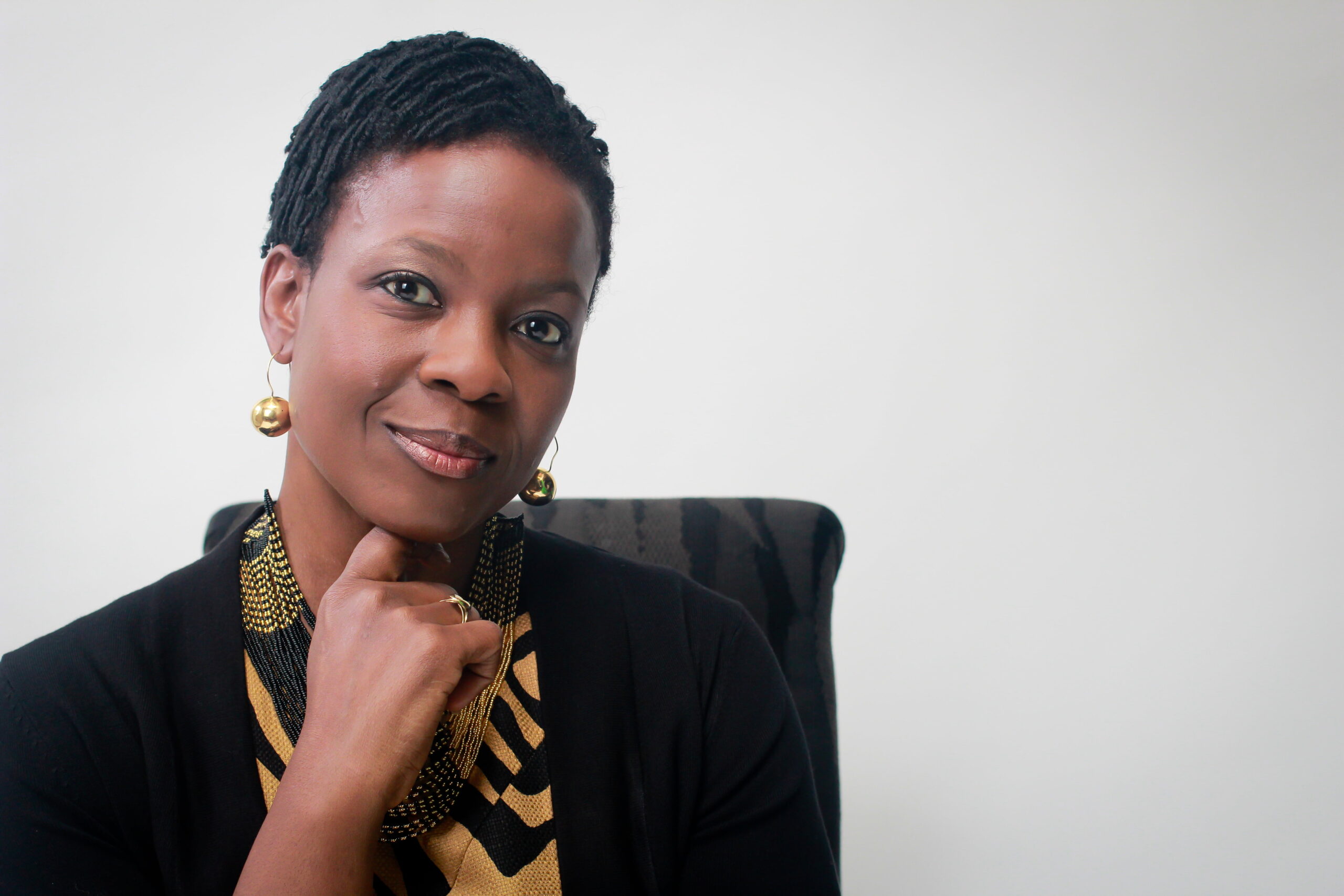 A person with short hair is sitting and smiling slightly, resting their chin on their hand. They are wearing a patterned top with a black cardigan and gold earrings. The background is plain and light-colored.