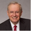 A smiling person with short gray hair and glasses is wearing a dark suit, white shirt, and red tie against a plain gray background.