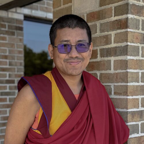 A person with short dark hair and glasses wearing a maroon and yellow robe, standing in front of a brick wall. They are smiling slightly and have their arms folded behind their back.