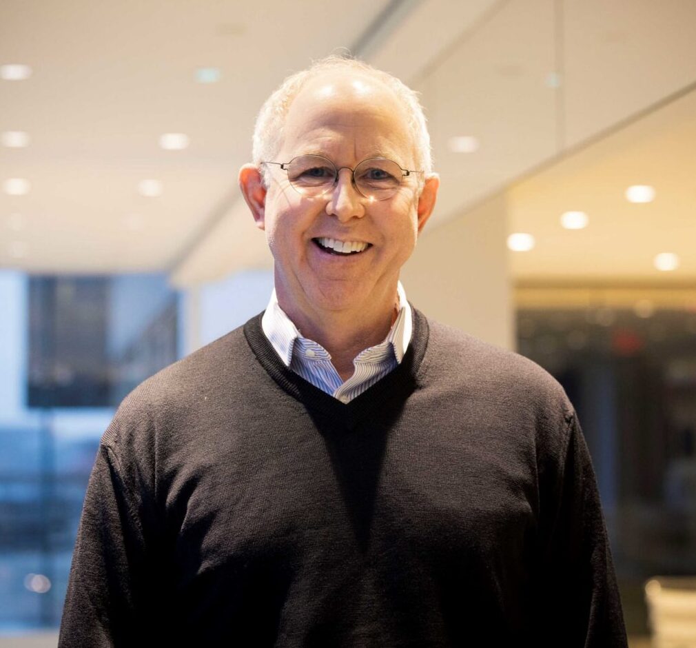 An older man with short gray hair, glasses, and a black sweater smiles while standing in a modern, well-lit office hallway with glass walls and ceiling lights.