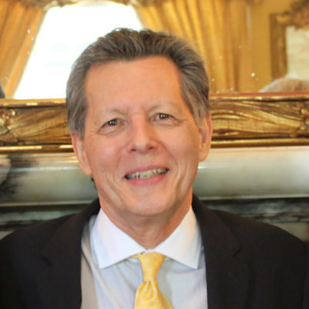 A man wearing a suit with a yellow tie is smiling. He stands in a room with elegant decor, featuring a large mirror and golden curtains in the background.