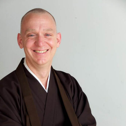 A smiling person with a shaved head wears a dark traditional robe with a white collar. The background is plain and light-colored. The individual appears calm and content, with their arms crossed and looking directly at the camera.