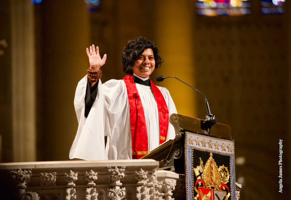 A smiling clergy member in white and red robes stands at a pulpit, raising one hand in greeting while speaking into a microphone in a large church.