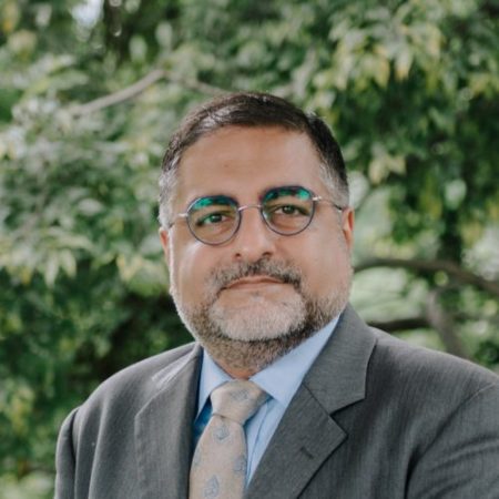 A man with a beard wearing glasses, a light blue shirt, a patterned tie, and a gray suit jacket. He is standing outdoors in front of a background with green, leafy trees.