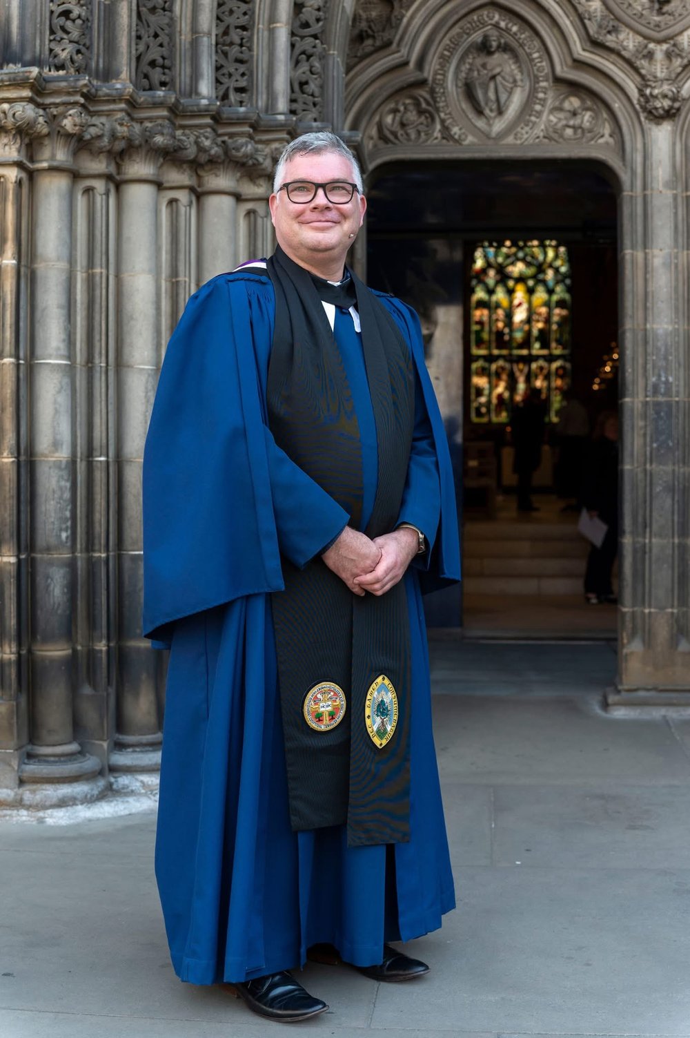 A smiling person in blue academic or clerical robes stands outside an ornate stone building with arched doorway and stained glass visible inside.