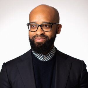 A bearded man wearing glasses and a dark suit with a striped shirt underneath stands against a white background, looking to the side with a slight smile.