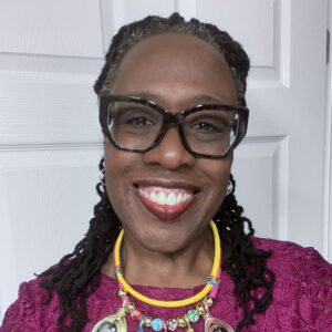 A person smiling at the camera, wearing glasses, a purple lace top, and a statement necklace with colorful beads and pendants. They are standing in front of a white door.
