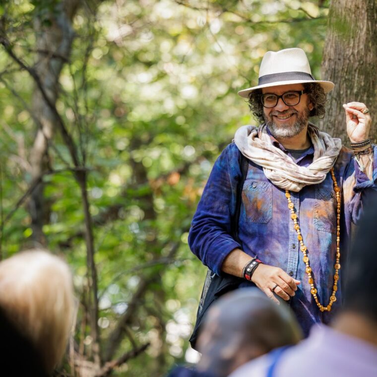 A man wearing a hat, scarf, glasses, and beaded necklaces smiles and gestures while standing among trees, addressing a group of people outdoors.