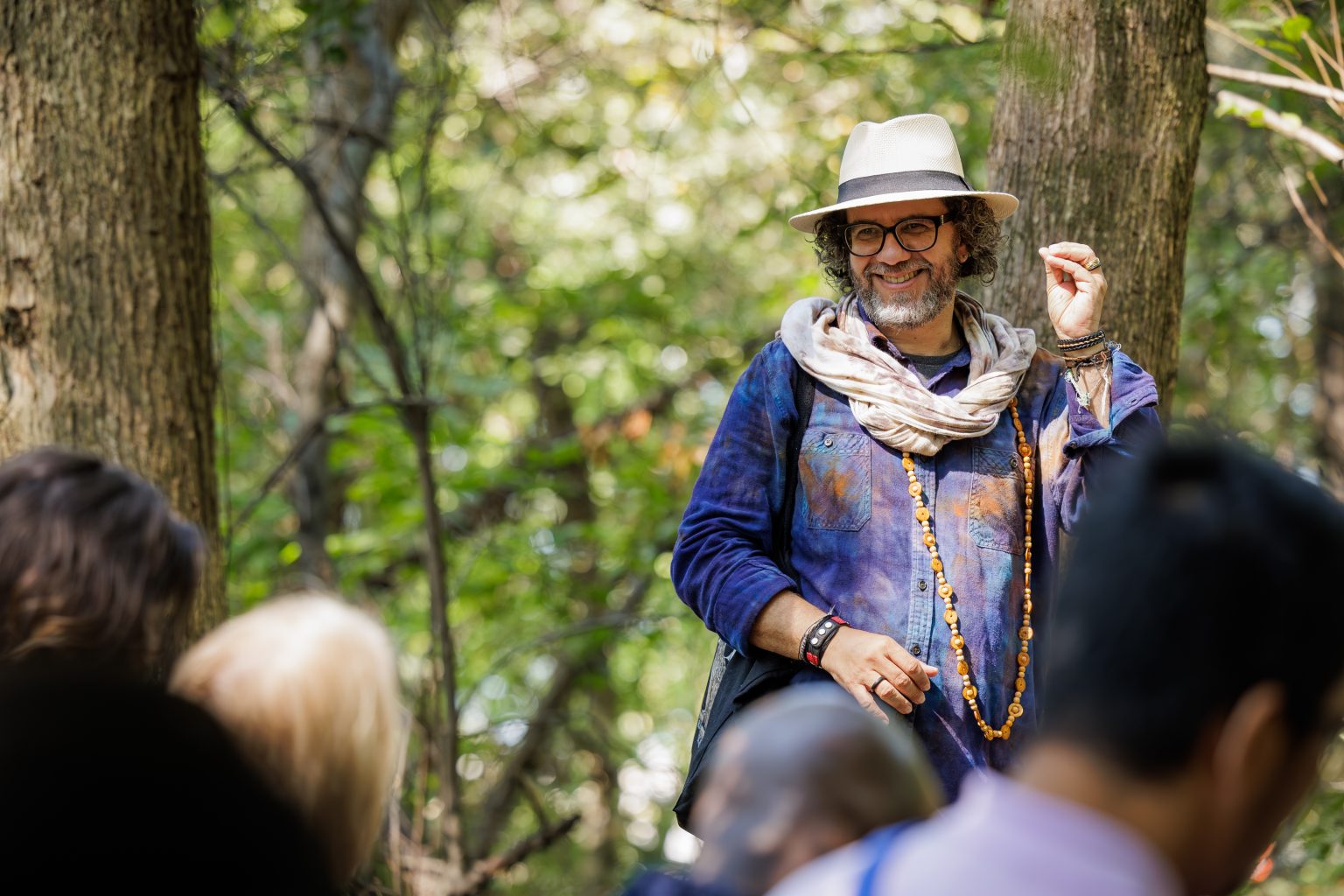 A man wearing a hat, scarf, glasses, and beaded necklaces smiles and gestures while standing among trees, addressing a group of people outdoors.