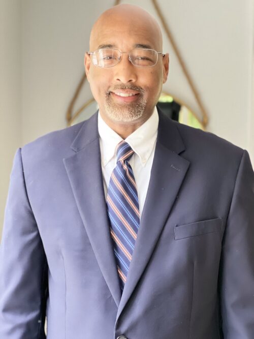 A man in a navy blue suit with a white shirt and striped tie stands smiling against a neutral background. He has a bald head, wears glasses, and has a neatly trimmed beard. The background is simple, with a glimpse of a mirror and a wooden object.