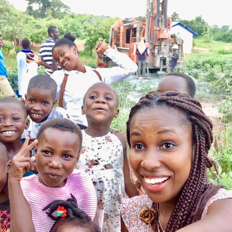 A joyful group of children and a young woman take a selfie outdoors. Several children smile and make playful gestures while trees and a drilling rig are visible in the background. The setting appears rural, with lush greenery surrounding them.