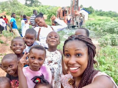 A joyful group of children and a young woman take a selfie outdoors. Several children smile and make playful gestures while trees and a drilling rig are visible in the background. The setting appears rural, with lush greenery surrounding them.