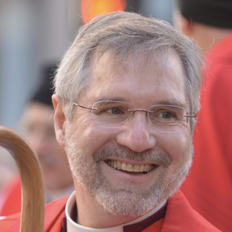 A smiling middle-aged man with gray hair, a beard, and glasses, wearing a red robe, is holding a wooden staff. The background is blurred, with other people dressed in similar red robes.