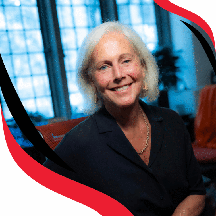 A smiling older woman with short white hair sits indoors near large windows, wearing a dark blouse and gold necklace. The photo has a red and black graphic border.