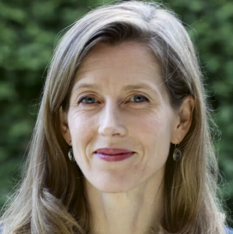 A woman with long, light brown hair smiles softly at the camera. She wears drop earrings and a light top. The background is blurred greenery.