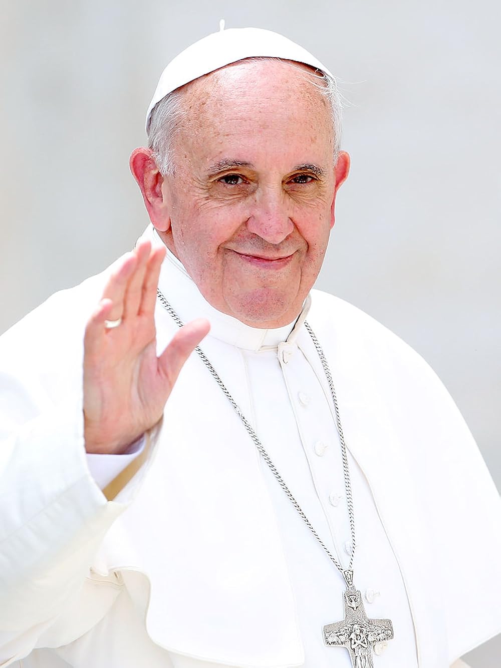 A man in white religious robes and a skullcap, resembling Pope Francis, waves and smiles at the camera with a cross necklace. The background is light and blurred.