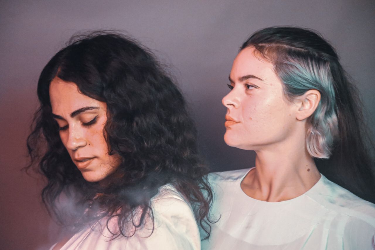 Two women in white tops stand back to back. One looks down with eyes closed and curly hair; the other faces sideways, gazing ahead with straight hair that has a light-colored streak. The background is gray with a soft, hazy effect.