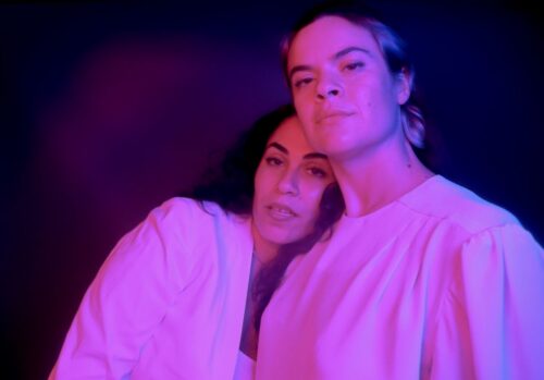 Two people in white tops pose closely together under purple and pink lighting, with one resting their head on the other’s shoulder, creating a soft, intimate atmosphere.
