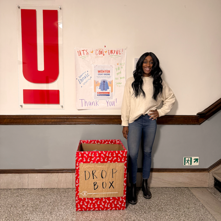 A person stands smiling beside a red donation box labeled "DROP BOX." Behind them is a poster for a winter coat drive and a large red "Y" symbol on the wall. The setting appears to be indoors with tiled flooring.