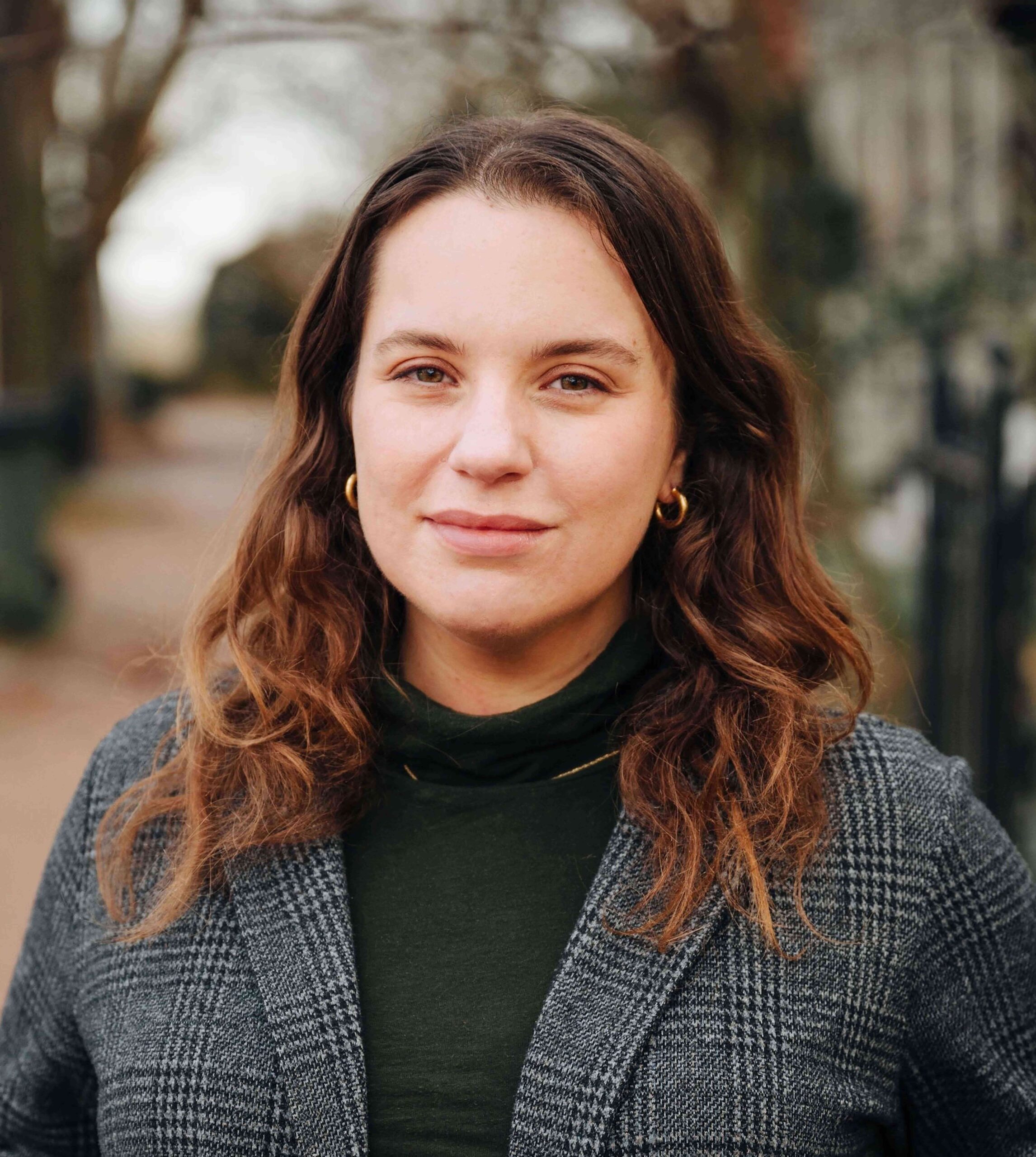 A woman with wavy brown hair wearing gold hoop earrings, a green turtleneck, and a gray plaid coat stands outdoors on a blurred, tree-lined path, looking calmly at the camera.