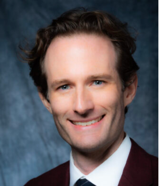 A man with light skin and short, wavy brown hair smiles at the camera. He is wearing a burgundy jacket, a white dress shirt, and a navy blue tie. The background is a dark, blurred gradient.