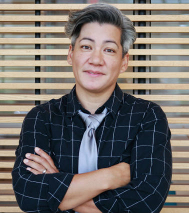 A person with short gray hair, wearing a black checkered shirt and a loose silver tie, is standing with their arms crossed in front of a wooden slatted background, smiling softly at the camera.