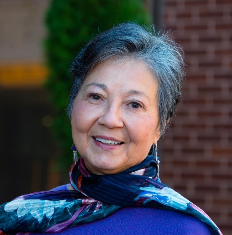 An older woman with short gray hair smiles gently at the camera. She wears a purple top and a colorful scarf, standing outdoors with a blurred background of greenery and brick.