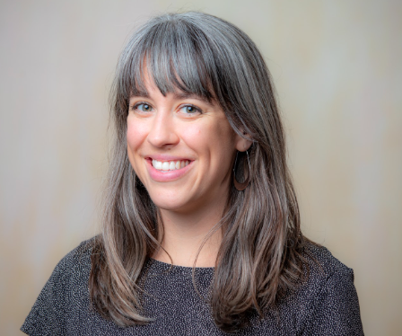 A woman with long, straight gray hair and bangs smiles at the camera. She is wearing a dark patterned top and hoop earrings, with a neutral, softly blurred background.
