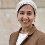 A woman wearing a beige headscarf, brown blazer, and white top smiles at the camera while standing in front of a light-colored stone wall.
