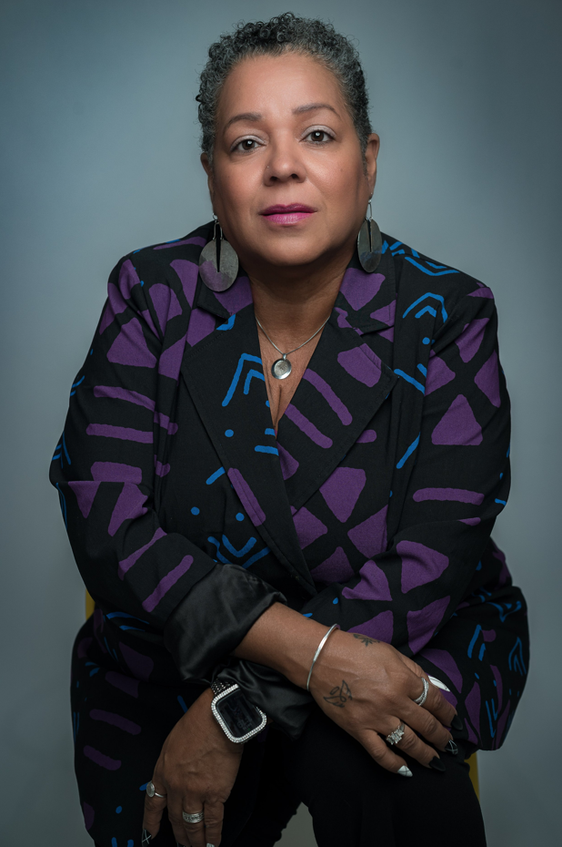 A confident woman with short curly gray hair sits with arms folded, wearing a bold black jacket with purple and blue patterns, silver jewelry, and a thoughtful expression on a plain gray background.