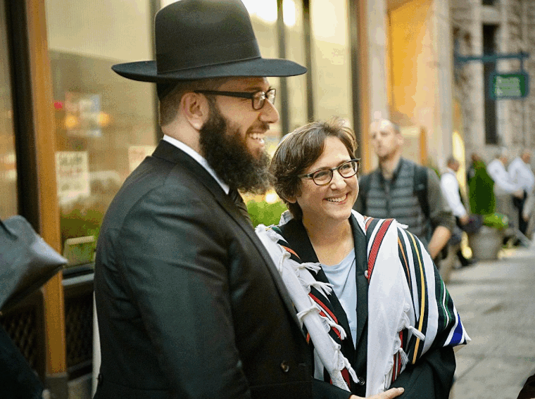 A man in a black hat and suit stands smiling next to a woman wearing glasses and a striped tallit. They are outside on a city street, surrounded by other people and buildings.