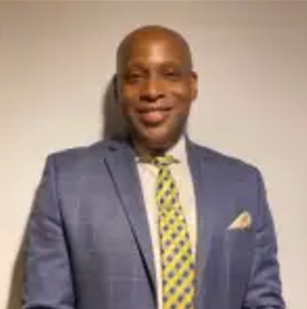 A man wearing a blue suit, light-colored shirt, and a yellow patterned tie stands against a plain light background, smiling at the camera.