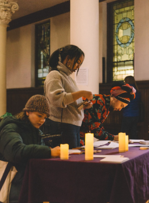 Three people are writing or signing papers on a purple table with lit candles, inside a room with stained glass windows and wooden walls.