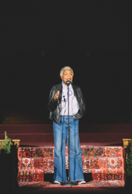 A person with short gray hair stands on a patterned carpeted stage, speaking into a microphone. They wear a black leather jacket, light blue shirt, and blue jeans. The background is dark and out of focus.