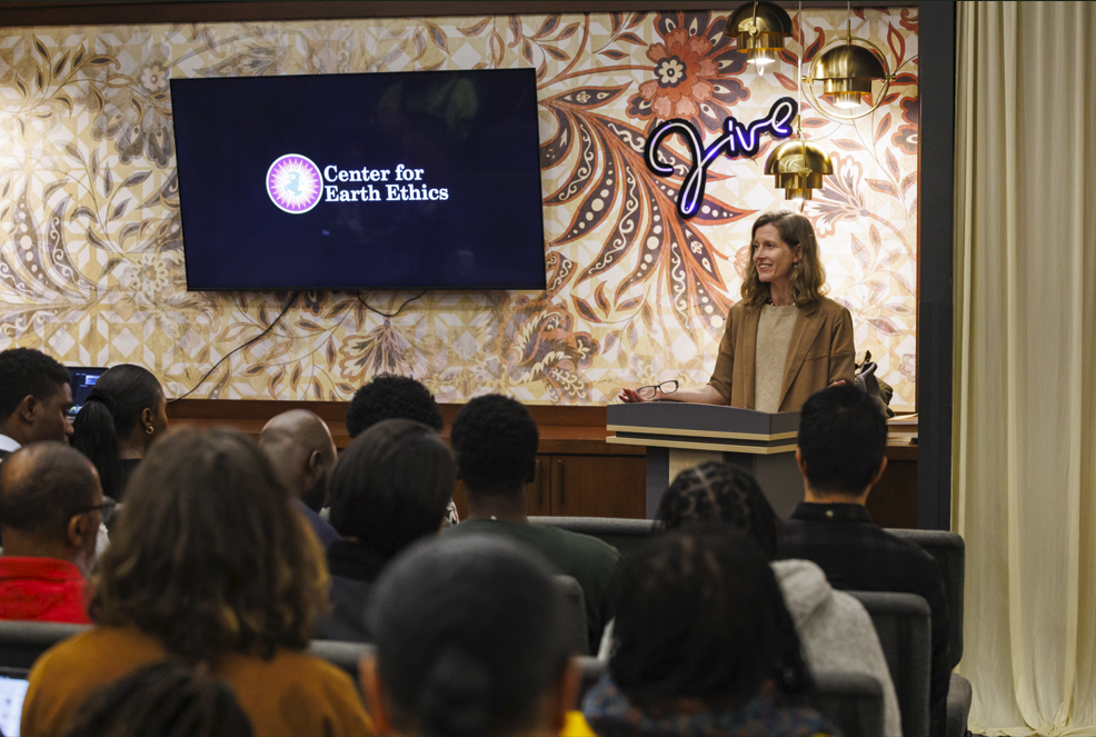 A woman speaks at a podium to an audience. Behind her is a screen displaying the "Center for Earth Ethics" logo and a neon sign reading "Live" on a decorative wall.