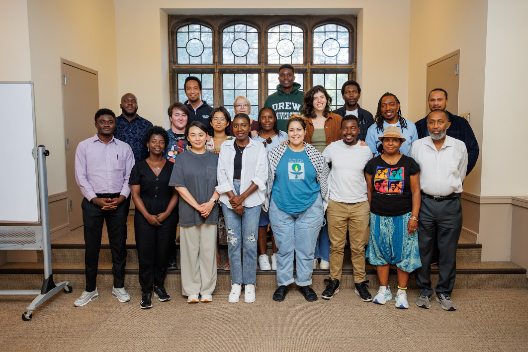 A diverse group of 19 people stands together indoors, smiling at the camera in two rows, with a large window in the background and a whiteboard to the left.
