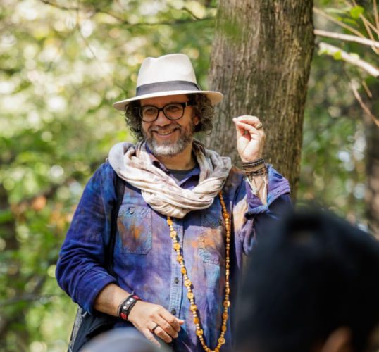 A smiling man with glasses, a white hat, and a scarf stands outdoors near a tree, wearing a blue shirt and a long beaded necklace, with greenery in the background.