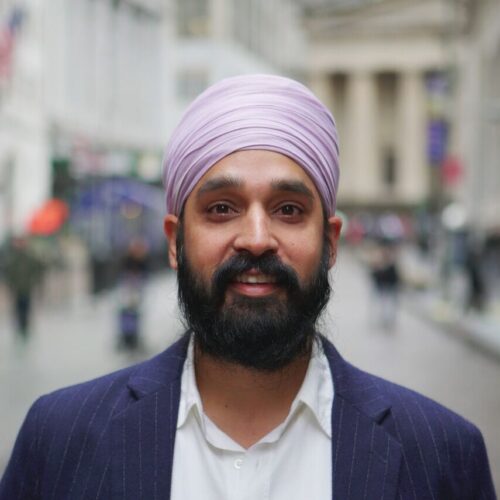 A person wearing a lavender turban and a dark striped blazer smiles while standing on a street with blurred buildings and pedestrians in the background.