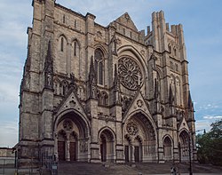A large stone Gothic-style cathedral with three arched doorways, pointed spires, and a prominent circular rose window above the main entrance, set against a blue sky.